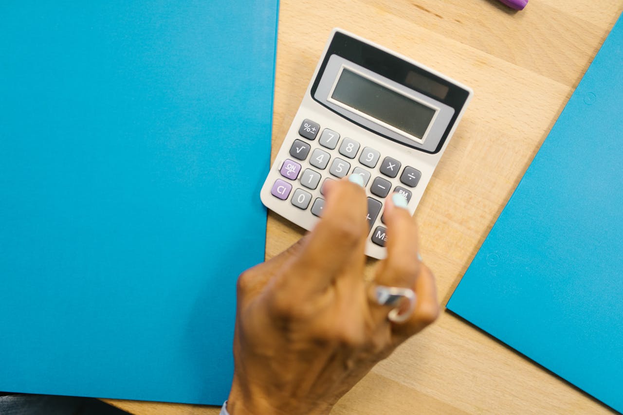 A hand using a calculator on a wooden desk surrounded by blue folders.