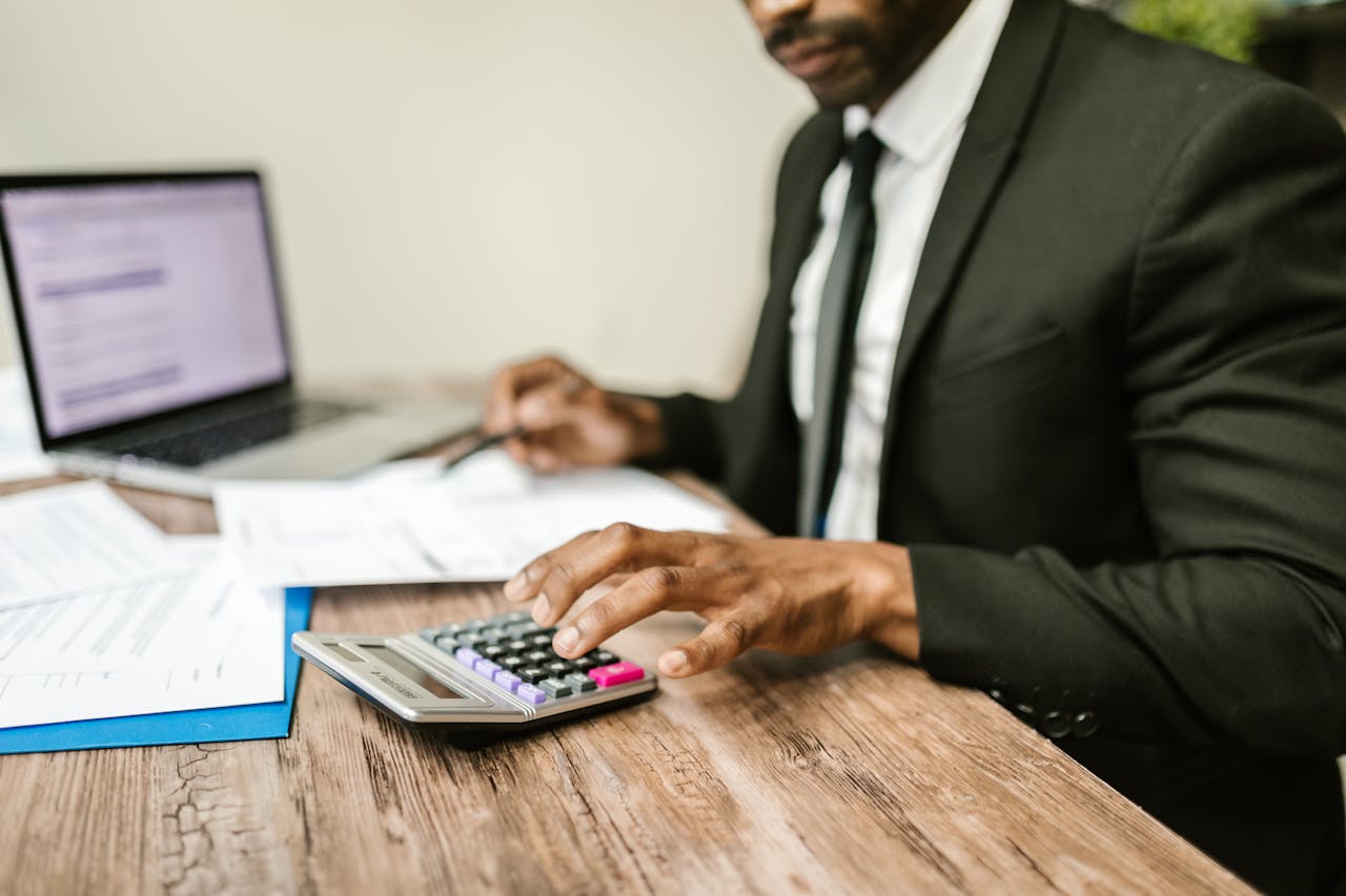 A businessman in a black suit using a calculator at a desk with financial documents and a laptop.